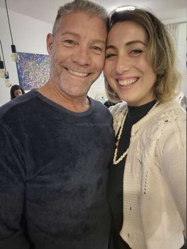 A smiling man and woman pose closely for a selfie in a cozy, well-lit room, radiating warmth and happiness. Colorful artwork decorates the background wall.