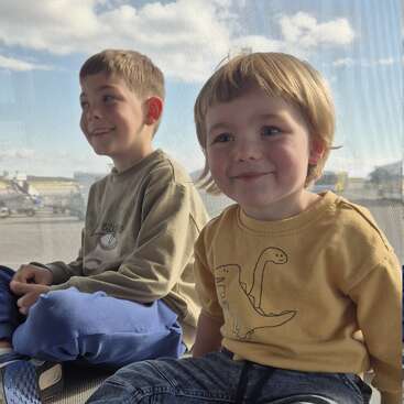 Two young children sit together, smiling and looking cheerful. Sunlight streams through the window behind them. They appear to be at an airport, waiting for a flight.