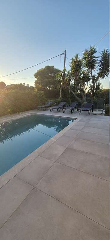 Une scène paisible autour d'une piscine extérieure au coucher du soleil, avec plusieurs chaises longues, des palmiers, un sol carrelé, une eau limpide, une végétation luxuriante et un ciel bleu serein.
