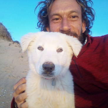 La imagen muestra a un hombre de pelo largo y camisa roja abrazando a un perro blanco en una playa de arena, con un cielo azul despejado.