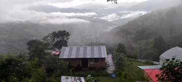 Una apacible casa está enclavada en exuberantes colinas verdes, rodeada de árboles y montañas neblinosas. Unas nubes bajas recorren el paisaje sereno y pintoresco del valle.