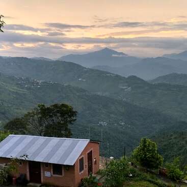A small house with a tin roof sits amongst lush greenery, overlooking rolling hills and mountains under a cloudy, golden sunrise sky, creating a serene landscape.