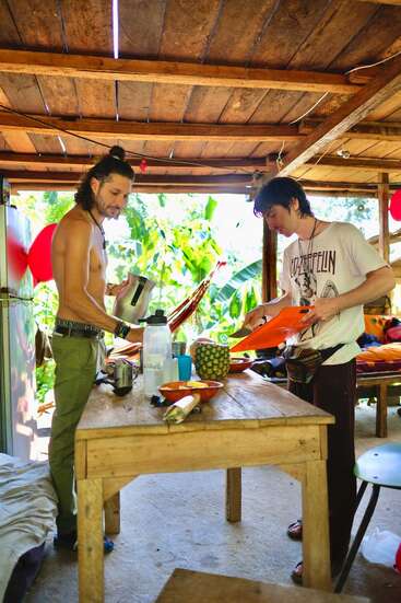 Deux hommes préparent ensemble des repas dans une cuisine rustique en plein air. L'un se sert dans une bouilloire, l'autre coupe des fruits en tranches. À l'extérieur, on aperçoit des feuillages tropicaux et des hamacs.
