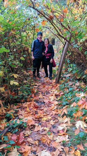 Duas pessoas sorriem em um caminho na floresta coberto de folhas coloridas de outono, cercadas por galhos arqueados e folhagens verdes e laranjas vibrantes, desfrutando de uma caminhada na natureza.