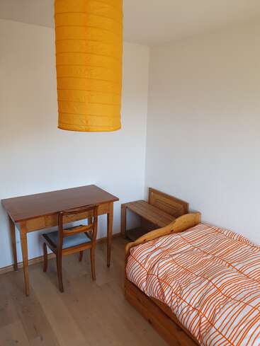 This image shows a simple, tidy room with wooden furniture: a desk, chair, small bench, and single bed with orange-striped bedding, under a large orange lampshade.
