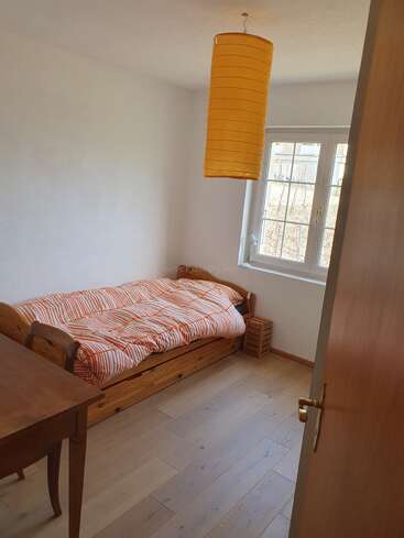 This is a small, simply furnished bedroom with a single bed, striped bedding, wooden furniture, a window, and a yellow cylindrical ceiling lamp. Cozy and bright.