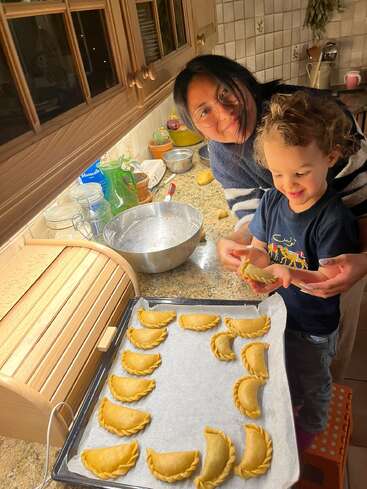 A woman and a child are happily making empanadas in a cozy kitchen. They smile while placing uncooked empanadas onto a baking tray lined with parchment paper.