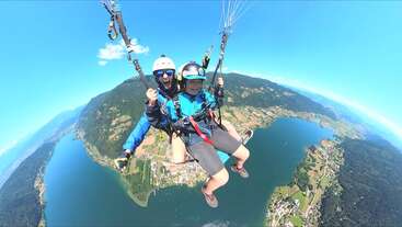 Zwei Menschen fliegen mit dem Gleitschirm hoch über einer wunderschönen Landschaft mit Seen, Bergen und Dörfern und genießen ein aufregendes Abenteuer unter einem klaren blauen Himmel. Lächeln und Aufregung!