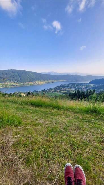 Eine Person in roten Schuhen entspannt sich auf einem grasbewachsenen Hügel und blickt auf einen ruhigen blauen See, ein entferntes Dorf, grüne Hügel und einen weiten blauen Himmel mit Wolken.