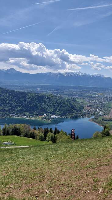 Eine atemberaubende Landschaft mit einem ruhigen See, umgeben von üppigen grünen Wäldern, sanften Hügeln und entfernten schneebedeckten Bergen unter einem strahlend blauen Himmel mit Wolken.
