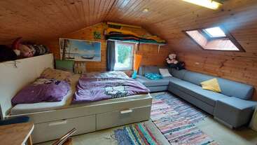 Dieses gemütliche Schlafzimmer im Dachgeschoss hat Holzwände, ein großes graues Sofa, ein Bett mit lila Bettwäsche, Stofftiere, einen bunten Teppich und Oberlichter.