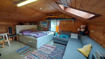 Dieses gemütliche Zimmer im Dachgeschoss hat holzgetäfelte Wände, ein großes Dachfenster, ein Bett, ein graues Sofa, bunte Teppiche und ein paar Stofftiere zur Dekoration.