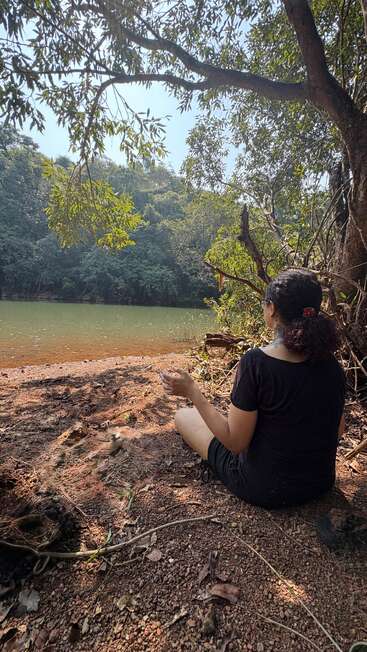 Uma mulher senta-se no chão perto de um lago sereno, cercada por árvores e pela natureza, desfrutando de um momento pacífico e contemplativo na sombra.