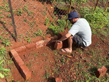 Um homem usando chapéu e chinelos está agachado, arrumando tijolos no solo vermelho perto de uma cerca de arame, provavelmente construindo um pequeno canteiro de jardim.