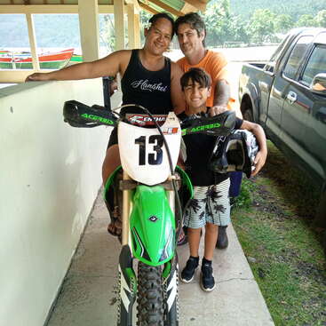 The image shows a family of three posing with a green and white dirt bike on a concrete porch, with a truck and boat visible in the background.