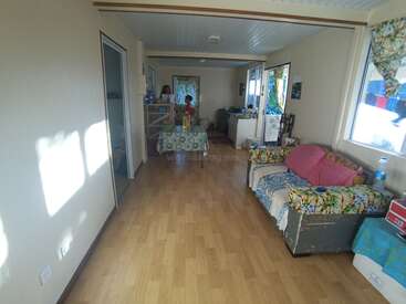 The image depicts a spacious room with a couch, table, and kitchen area, featuring white walls and a light-colored wood floor, bathed in natural sunlight.