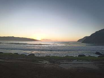 The image depicts a serene beach scene at sunset, featuring a tranquil ocean, a rocky shoreline, and a majestic mountain range in the distance, with a clear sky above.
