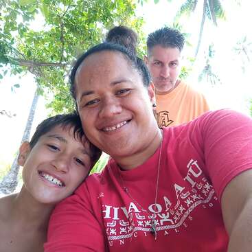 The image shows a woman and a boy posing for a selfie, with a man standing behind them in a tropical setting.