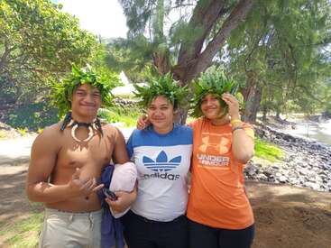 The image depicts three individuals standing together, adorned with green leafy headpieces, posing for a photo in a tropical setting with trees and a body of water in the background.