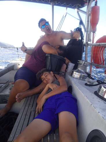 The image depicts a man and a boy on a boat, with the man holding a bucket and the boy lying down, both appearing to be in a relaxed state.