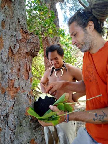 The image depicts two men standing by a tree, with one holding a black object and the other wearing a necklace made of animal teeth.