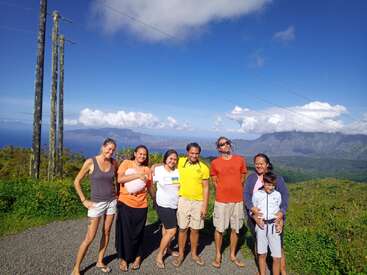 The image depicts a group of seven people posing for a photo on a gravel road, set against a picturesque backdrop of mountains and a blue sky with clouds.