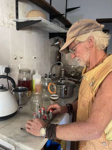 An older man with white hair and glasses works in a kitchen, adjusting a device on the counter. There are jars, a kettle, and utensils nearby.