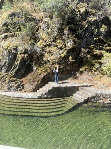 A person stands on stone steps beside a clear, green pool of water, surrounded by rocky cliffs covered in moss and vegetation under bright sunlight.
