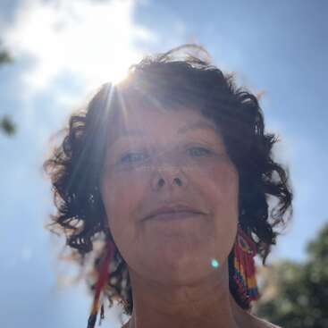 Una mujer con el pelo rizado y pendientes de cuentas está de pie al aire libre. El sol brilla intensamente detrás de su cabeza, creando un efecto de halo radiante. Se ven el cielo y los árboles.