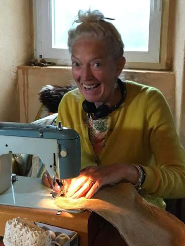 A cheerful woman with blonde hair, wearing a yellow sweater, smiles widely while sewing fabric with a vintage sewing machine next to a window. Cozy setting.