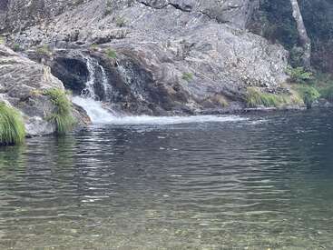 Ein kleiner Wasserfall stürzt über zerklüftete Felsen in einen ruhigen, klaren Pool, der von saftig grünem Gras und einer friedlichen Naturlandschaft umgeben ist und zu Entspannung und Ruhe einlädt.