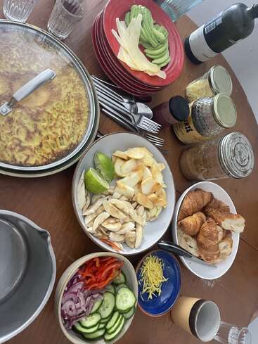 Un delicioso brunch con croissants, rodajas de verduras, aguacate, queso, pollo, condimentos, una gran frittata, vasos, platos y una botella de vino.