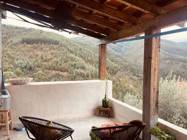 Una acogedora terraza cubierta con vistas a exuberantes montañas verdes. Unas sillas sencillas, una mesita con una botella de cristal verde y unas rústicas vigas de madera crean un ambiente relajante y acogedor.