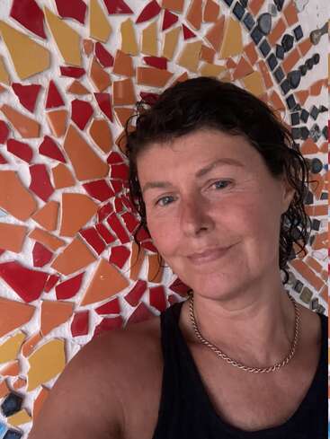 Une femme aux cheveux courts, noirs et mouillés sourit doucement. Elle porte un collier en chaîne en or et un haut noir, sur fond de mosaïque aux couleurs vives.