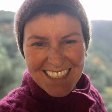 Eine Frau, die einen kastanienbraunen Pullover und eine braune Mütze trägt, lächelt strahlend in die Kamera, während der natürliche Hintergrund im Freien verschwommen ist.