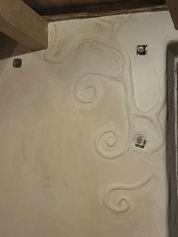 A imagem mostra uma parede de gesso com padrões artísticos em espiral esculpidos na superfície. Vêem-se vigas de madeira e caixas elétricas à mostra, o que dá um ar rústico e de obra em andamento.