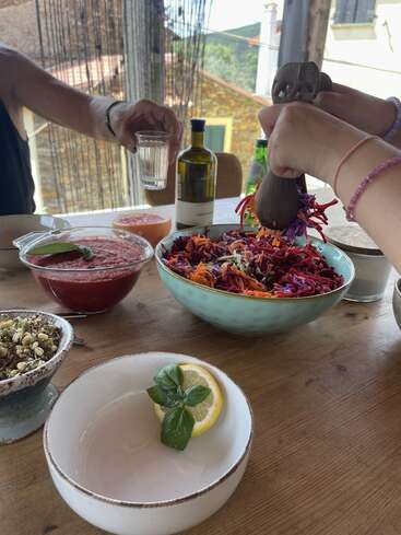 Uma mesa bem animada, com uma salada fresca e colorida, sopa e brotos. Mãos servindo a comida, um copo de água erguido, limão, uma guarnição de manjericão e azeite.