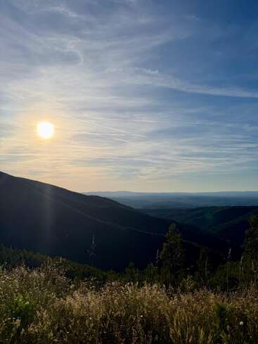 A luz dourada do sol banha as colinas onduladas e os prados verdejantes, projetando longas sombras. Nuvens suaves e rastros de avião se estendem por um céu azul sereno acima da paisagem pitoresca.