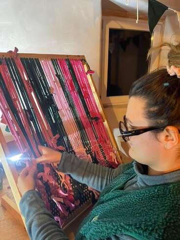 Uma mulher de óculos tece cuidadosamente fios coloridos em tons de rosa e verde num tear de madeira, concentrada no seu projeto têxtil criativo, numa sala aconchegante.