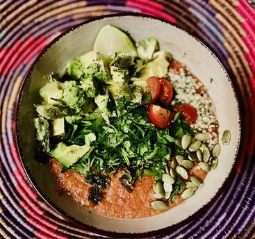 Uma tigela cheia de cor com abacate cremoso, tomates-cereja, sementes, ervas e limão sobre uma base rica e espessa, disposta sobre um fundo colorido de esteira trançada. Uma delícia saudável.