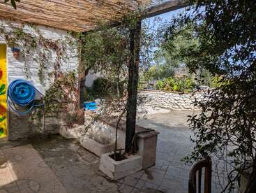 Um pátio ensolarado com plantas trepadeiras, uma mangueira de jardim azul, mural amarelo, piso de ladrilho, caixas de plantas de pedra, telhado de bambu e uma parede de pedra com vegetação.