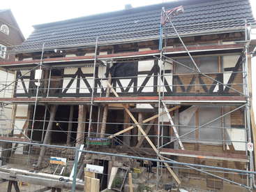 L'image représente une maison en construction, avec des échafaudages et des poutres en bois apparentes, avec un toit en tuiles et un ciel nuageux en arrière-plan.