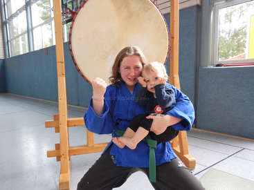 L'image montre une femme en uniforme bleu d'arts martiaux tenant un bébé en uniforme noir, avec un grand tambour derrière elles dans un gymnase.
