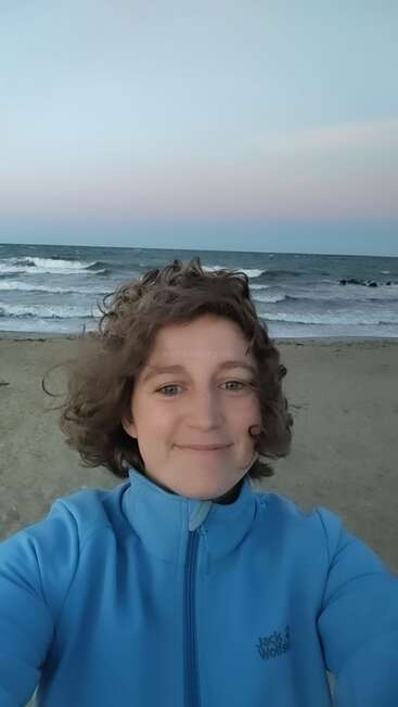 Une personne aux cheveux bouclés sourit, vêtue d'une veste bleu clair, sur une plage de sable. Derrière eux, les vagues s'écrasent sous un ciel crépusculaire aux couleurs pastel.