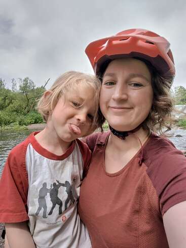Une femme portant un casque rouge sourit en prenant un selfie avec un jeune enfant qui tire la langue de manière ludique. Ils sont en plein air, près de l'eau et des arbres.