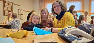 Une salle de classe remplie d'enfants et d'adultes s'adonnant à des activités artistiques et manuelles. Trois personnes sourient à la caméra, entourées de bateaux en papier colorés posés sur la table.