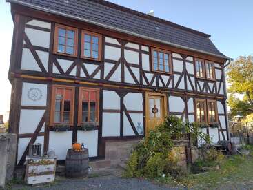 Cette image montre une charmante maison à colombages avec des poutres brunes et des panneaux blancs. Il y a des cadres de fenêtres orange, des tonneaux décoratifs, des plantes et une porte d'entrée accueillante.