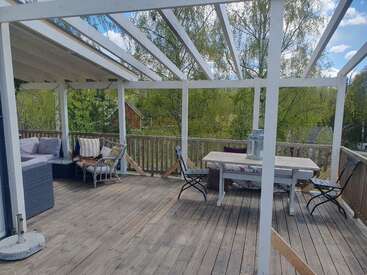 Das Bild zeigt eine Holzterrasse mit teilweiser Überdachung, Sitzgelegenheiten im Freien, darunter ein Sofa und Stühle, sowie einen Tisch, umgeben von grünen Bäumen und blauem Himmel.