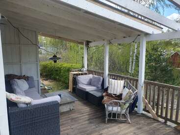 Eine gemütliche Terrasse mit gepolsterten Korbmöbeln, Kissen, einer Decke, Lichterketten, Holzterrasse, teilweisem Dach und umgeben von grünen Bäumen und Sträuchern.