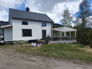 Ein weißes zweistöckiges Haus mit dunklem Dach, Holzterrasse und überdachter Pergola, umgeben von Bäumen und Rasen unter einem teilweise bewölkten Himmel. Draußen liegen Gartengeräte herum.